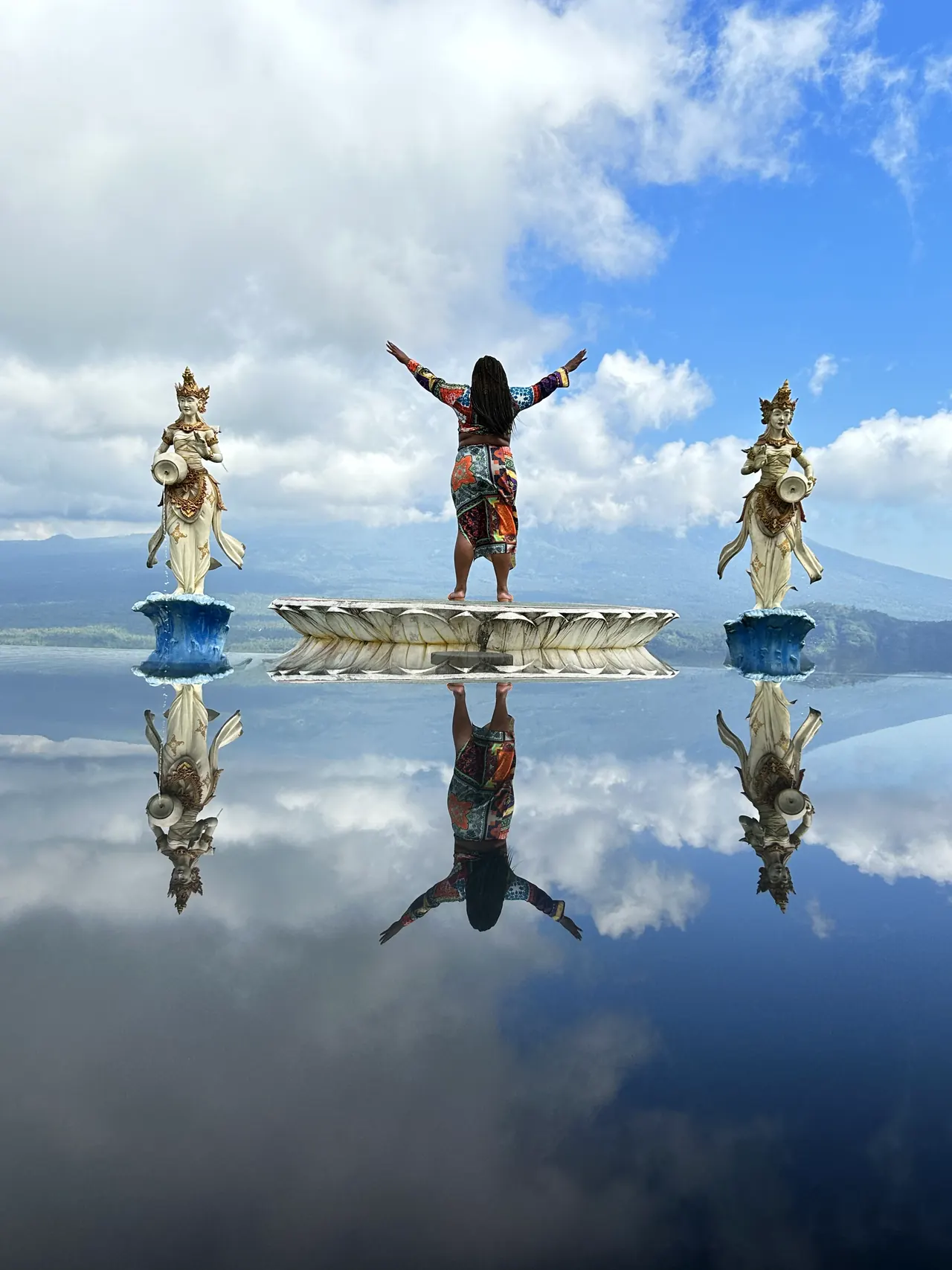 Statues and a visitor with raised arms reflected on a mirror-like water surface
