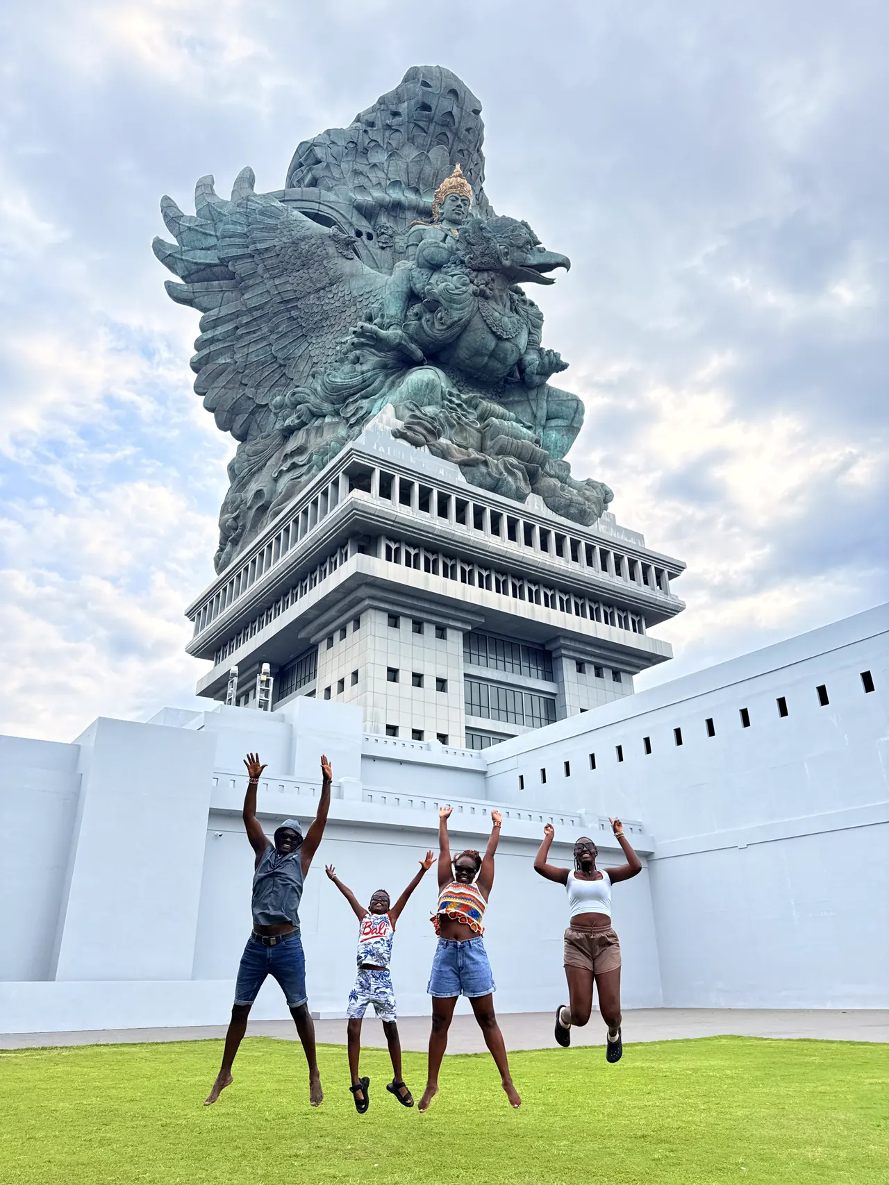 Four visitors leaping in front of the white base of the Garuda Wisnu Kencana statue